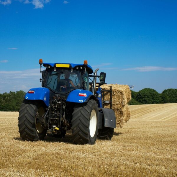 A blue tractor carries a large square hay bale across a cleared field under a bright blue sky.